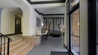 This single-floor space makes a strong first impression upon entry, where large glass-paneled doors open to a tiled foyer anchored by a welcome mat and flanked by exposed brick walls. A few steps lead up to the main interior, which transitions seamlessly between a dining area and a lounge space. The dining area features a sleek dark table paired with high-backed white chairs, while a contemporary wall sconce and abstract artwork contribute to the refined aesthetic.

The most striking feature of the space is a large-scale typographic mural adorning the far wall, displaying world city names — including Oslo, Nairobi, Warsaw, Reykjavik, Istanbul, and Zagreb — in bold black and white. This graphic element sets a cosmopolitan, design-forward tone throughout.

The lounge area includes a blue sofa, a dark coffee table, and a patterned area rug, creating a comfortable yet sophisticated seating arrangement. Dark crown molding frames the white walls and ceiling, adding architectural contrast and definition. The overall ambiance is modern and curated, with a cohesive monochromatic palette punctuated by carefully chosen design elements.