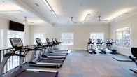 Located at 1910 Capital Creek Dr in Wake Forest, NC, this fitness center offers a clean, modern environment designed to support an active lifestyle. The single-floor facility is spacious and thoughtfully laid out, providing ample room for a full range of cardio equipment, including a row of treadmills and a dedicated section of stair climbers and elliptical machines — all from True Fitness, a well-regarded commercial brand. The flooring features a combination of neutral and blue-accented carpet tiles that define workout zones while adding a polished aesthetic to the space. The high coffered ceiling enhances the sense of openness and is equipped with multiple ceiling fans to ensure comfortable airflow throughout. Recessed lighting and sleek linear light fixtures provide consistent, even illumination across the entire floor. Multiple windows along the perimeter walls allow natural light to fill the room and offer views of the surrounding greenery, creating an inviting atmosphere. The neutral color palette of soft whites and warm beiges contributes to a calm and focused workout environment. Overall, this fitness center combines functional design with a bright, welcoming ambiance well-suited for daily exercise routines.