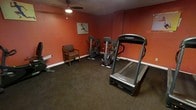 This fitness center offers a focused and functional workout environment designed to support cardio-based exercise routines. The room features bold, deep-red walls that create an energizing atmosphere, complemented by dark carpeted flooring and white baseboards that frame the space cleanly. A ceiling fan overhead provides air circulation, helping to keep the environment comfortable during workouts. The equipment lineup includes a treadmill prominently positioned for easy access, two stationary upright bikes, and an additional piece of cardio equipment along the far wall, offering users a variety of aerobic workout options. A cushioned armchair is available for rest between sets or for a companion waiting nearby. Motivational athletic artwork adorns the wall, adding to the active ambiance of the room. The layout is efficient and well-organized, maximizing the available floor space for user comfort and ease of movement between machines. This single-floor facility provides a convenient, no-frills fitness option with all the essential equipment needed for a productive cardio session.