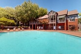 Located at 4659 Dawson Blvd in Atlanta, Georgia, this outdoor amenity area is centered around a generously sized swimming pool with clear turquoise water. The pool is surrounded by a well-maintained brick paver deck that provides ample space for lounging and socializing. Multiple chaise lounge chairs and patio seating with umbrella tables are arranged around the perimeter, offering comfortable options for relaxation under the open sky.

The area is fully enclosed by a sturdy wrought iron fence with stone pillar accents, providing both security and a refined aesthetic. Entry is controlled through gated access points, with keypad or card-access hardware visible at the entrance. Mature trees surround the space, casting natural shade and creating a private, park-like atmosphere that offers a retreat from the surrounding urban environment.

The multi-story building adjacent to the pool features a mix of brick and siding construction, with covered balconies and gabled rooflines that lend a traditional architectural character. A secondary upper-level deck overlooking the pool adds vertical dimension to the space. The combination of classic brick detailing, lush greenery, and well-appointed outdoor furnishings creates a welcoming and visually appealing communal environment suited for warm Georgia days.