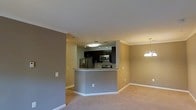 This single-floor space showcases an open-concept design that seamlessly connects the kitchen and living area, creating a cohesive and functional layout. The kitchen is equipped with stainless steel appliances, including a refrigerator, microwave, and range, all framed by dark espresso cabinetry that contrasts elegantly with the granite-topped breakfast bar. The bar serves as both a workspace and a casual dining area, forming a natural boundary between the kitchen and the adjacent living space. Track lighting illuminates the kitchen, while a decorative chandelier anchors the dining or living zone, adding a touch of warmth to the space. Neutral-toned walls in a warm taupe complement the light beige carpeting that flows throughout the living area, contributing to a clean and cohesive aesthetic. Crown molding runs along the ceiling, lending a refined architectural detail to the interior. The layout maximizes the sense of openness, making the space feel well-proportioned and inviting. White baseboards provide crisp contrast against the walls and flooring, reinforcing the overall polished, move-in-ready character of the space.