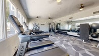 Located on E Fifth Street in Tucson, Arizona, this fitness center offers a well-organized and fully equipped workout space designed to support a comprehensive exercise routine. The room is contained on a single level and provides a comfortable sense of openness, aided by neutral light-toned walls and recessed lighting supplemented by ceiling fans that promote airflow throughout the space.

The flooring features a distinctive geometric-patterned carpet in varying shades of gray, lending the room a modern, dynamic aesthetic. Along one wall, a bank of windows allows natural light to filter in, offering views of the surrounding outdoor environment including mature palm trees. A large mirrored wall on the opposite side visually expands the space while allowing users to monitor their form during workouts.

Equipment is thoughtfully arranged throughout the room. Cardio machines, including treadmills, ellipticals, and stair climbers, are positioned near the windows. A cable machine system, adjustable weight bench, and a full rack of dumbbells provide ample strength-training options. Medicine balls and additional accessories are visible along the mirrored wall. A wall-mounted flat-screen television adds entertainment, while motivational phrases such as "Don't Wish For It, Work For It" and "Beast Mode On" are displayed prominently, contributing to an energizing atmosphere.