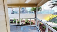 This single-floor space features a covered outdoor patio that serves as a welcoming transitional area between the interior and the surrounding grounds. The patio is enclosed by clean white railings and benefits from a protective overhead covering, making it suitable for year-round use regardless of weather conditions. Overhead fluorescent lighting ensures the space remains well-lit even when natural light is limited.

Directly adjacent to the patio, neatly maintained landscaping beds are filled with mulch and an assortment of rounded shrubs and green plantings, adding a fresh and tidy aesthetic to the outdoor area. A prominent palm tree rises just beyond the railing, lending a warm, subtropical character to the environment.

The patio floor is a smooth concrete surface, offering a durable and easy-to-maintain foundation. Looking outward, views extend to the wider community, including additional buildings, staircases, and open communal spaces enclosed by fencing. A security camera is mounted on the overhead structure, indicating a focus on safety throughout the property. The overall ambiance is bright, open, and well-kept, making this covered patio a pleasant outdoor retreat within a larger residential or mixed-use community setting.