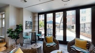 Located at 600 St. Mary's St in Raleigh, NC, this single-floor amenity lounge offers a thoughtfully designed communal space with a coastal-inspired aesthetic. The interior is anchored by a striking wood-paneled media wall, against which teal cabinetry and a wall-mounted flat-screen TV create a vibrant focal point. An elegant orb chandelier overhead adds a sophisticated touch to the seating area, which is furnished with woven lounge chairs accented by bold yellow and patterned cushions, low round coffee tables, and a neutral sectional sofa.

A standout feature is the dramatic blue watercolor accent wall, adorned with textured woven artwork, which adds depth and artistic character to one end of the room. Built-in shelving along the opposite wall provides additional display and storage. Wide-plank wood-look flooring runs throughout, complementing the light, neutral palette.

Floor-to-ceiling dark-framed glass doors and windows flood the space with abundant natural light and provide direct views of and access to an outdoor courtyard featuring shaded seating, a pool area, and surrounding brick architecture. A bistro-height table with teal barstools near the window wall offers an additional casual seating option. A glass-panel door leads to an adjacent room, rounding out this well-appointed and inviting community space.
