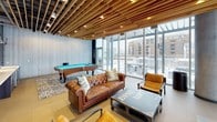Located at 1155 N Howe St in Chicago's vibrant Old Town neighborhood, Esprit Loft is a thoughtfully designed amenity lounge that blends industrial sophistication with warm, inviting elements. The space is anchored by a striking slatted wood ceiling installation that adds warmth and architectural depth, complementing the exposed concrete columns and sleek grey-toned walls throughout.

Floor-to-ceiling glass walls line the perimeter, flooding the interior with abundant natural light and offering expansive views of the Chicago skyline and adjacent outdoor terrace. The open layout is organized into distinct functional zones, beginning with a comfortable lounge area furnished with tufted brown leather Chesterfield sofas, eclectic accent chairs in mustard and teal, and an industrial-style coffee table bearing the Esprit Loft branding.

A full-size pool table set over a bold patterned area rug serves as a central entertainment feature. Adjacent to it, a built-in wet bar with cabinetry, a beverage refrigerator, and a wall-mounted flat-screen television creates an ideal social hub. A glass-enclosed private room provides a quieter retreat within the larger space, while pendant lighting adds a refined finishing touch throughout. The overall ambiance is modern, energetic, and perfectly suited for social gatherings.