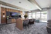 This contemporary amenity lounge is thoughtfully designed to accommodate both collaborative work and casual socializing. The space is anchored by a substantial wooden conference table surrounded by woven upholstered chairs, providing an inviting setting for meetings or group gatherings. Exposed rustic wood beams run across the ceiling, adding warmth and architectural character that contrasts beautifully with the clean, modern finishes throughout.

Large floor-to-ceiling windows and sliding glass doors flood the space with natural light while offering views of the surrounding outdoor courtyard and neighborhood. A built-in window seat with decorative pillows provides an additional spot for relaxed seating. Patterned carpeting in cool blue-gray tones adds texture underfoot and complements the neutral color palette of the walls and furnishings.

Along one wall, natural wood paneling paired with a herringbone tile accent backdrop frames a flat-screen television and a credenza with storage. A secondary lounge area with a long counter workspace lines another wall, accompanied by slim upholstered chairs — well suited for individual work or study. A staircase with warm wood treads, a black metal railing, and decorative hexagonal wall art connects to an adjacent level. Together, these elements create a polished, multi-functional space with a welcoming, modern-rustic ambiance.