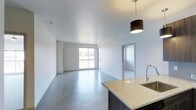 This contemporary single-floor space showcases a clean, modern aesthetic defined by an open-concept floor plan and a bright, neutral palette throughout. The polished concrete floors lend an industrial-chic character while offering exceptional durability and ease of maintenance. The kitchen features a sleek dark-toned island topped with speckled granite-style countertops, a stainless steel sink, and a stylish cylindrical pendant light overhead, creating a functional yet visually striking focal point. The open living area flows seamlessly from the kitchen, bathed in natural light streaming through a large sliding glass door that opens onto a private balcony with railing — ideal for bringing in fresh air and outdoor views. The expansive white walls and smooth ceilings amplify the sense of openness throughout the space. A separate doorway leads to an additional room, providing flexible use as a bedroom, office, or storage space. The minimalist design approach, combined with high-quality finishes and thoughtful layout, makes this a versatile and well-appointed space suited to a variety of lifestyle needs.