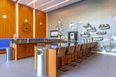This contemporary community clubhouse is designed to offer residents a sophisticated and comfortable gathering space. The open-plan layout encompasses a generously sized lounge area anchored by deep navy blue upholstered seating, including plush sofas and accent chairs arranged around a central entertainment wall fitted with multiple flat-screen displays. A striking cityscape-inspired mosaic tile feature wall serves as a dynamic artistic backdrop throughout the space.

The dedicated coffee bar is a standout amenity, equipped with a built-in coffee machine, dark granite countertops, warm wood cabinetry, open shelving stocked with décor and supplies, and a full sink — creating a fully functional refreshment station. Adjacent to the coffee bar, a dining area provides a glass-top table with woven chairs, ideal for communal meals or informal meetings.

Tall floor-to-ceiling windows flood portions of the space with natural light while offering direct views of the outdoor pool courtyard. A bright seating nook near the entry features a bold blue velvet sofa beneath a sculptural pendant light, framed by lush greenery. The ceiling design incorporates recessed linear lighting alongside dark structural beams, lending an industrial-modern aesthetic. Glass entry doors and large-format tile flooring complete the polished, contemporary character of this well-appointed communal facility.