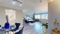 This thoughtfully designed single-floor residence offers an inviting open layout that seamlessly connects its key living spaces. The entryway greets you with warm wood-look flooring, crown molding, and a decorative console, setting an elegant tone throughout. Adjacent to the entry, a compact dining area with a bistro-style table flows naturally into the spacious living room, where a ceiling fan, large windows with shutters, and abundant natural light create a bright and airy atmosphere. The living room is anchored by a white sofa, a tufted royal blue ottoman, and striking blue velvet accent chairs, all complemented by bold abstract artwork and lush indoor plants.

The galley-style kitchen features grey shaker cabinetry, light quartz countertops, stainless steel appliances including a refrigerator and dishwasher, and tile flooring — offering a clean and functional workspace.

The bedroom is a tranquil retreat, appointed with plush carpeting, crown molding, a tufted navy headboard, and decorative wall art. A sliding glass door opens directly onto a private balcony that overlooks a sparkling pool and lush landscaping, providing a peaceful outdoor escape. A generous closet and additional seating area within the bedroom add further comfort and practicality to this well-appointed space.