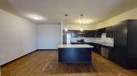 This residence at Springs At Hamburg presents a well-planned single-floor layout that balances modern aesthetics with everyday functionality. The kitchen serves as a standout centerpiece, featuring dark espresso cabinetry, granite-style countertops, a full suite of stainless steel appliances including a refrigerator, range, microwave, and dishwasher, and a built-in wine rack. A large central island with pendant lighting provides additional prep space and a casual dining option. Wood-style flooring extends throughout the kitchen and open living area, creating a warm and cohesive feel. The living space is generously proportioned, offering a bright and versatile area ideal for a variety of layouts. The bedrooms are carpeted for comfort and feature large windows with blinds that invite natural light and outdoor views. One bedroom is further enhanced with a ceiling fan. The bathrooms are finished with dark vanity cabinetry, granite-style countertops, wood-look flooring, and full-size soaking tubs with shower surrounds. The consistent use of dark cabinetry, neutral walls, and quality fixtures throughout the unit gives the space a cohesive, contemporary character well-suited for comfortable everyday living.