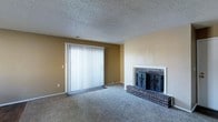 This single-floor residence offers a comfortable and functional layout designed for everyday living. The living area serves as the heart of the home, highlighted by a classic brick fireplace with elegant black metal doors — a standout architectural feature that adds warmth and character to the room. The fireplace is set against neutral-toned walls that create a calm, inviting atmosphere throughout the space. Plush wall-to-wall carpeting provides a soft underfoot feel, contributing to the overall coziness of the living area. A large sliding glass door with vertical blinds dominates one wall, allowing abundant natural light to pour into the room while offering potential access to an outdoor area. The textured popcorn ceiling adds a traditional residential charm consistent with the overall aesthetic of the home. The neutral color palette of warm beige walls and grey carpeting creates a versatile backdrop, making it easy to personalize with furnishings and décor. Overall, this single-story space combines classic design elements with practical features, offering a welcoming environment suitable for a variety of living arrangements.