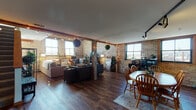 This two-floor loft-style space showcases a compelling blend of industrial heritage and warm, rustic character. The main level opens into a generously proportioned open-concept living, dining, and kitchen area anchored by striking exposed brick walls, dark hardwood floors, and structural timber posts that add architectural authenticity. Large multi-pane windows flood the living area with abundant natural light and frame outdoor views, while decorative string lights contribute a cozy, inviting ambiance. The living room offers ample space for multiple seating arrangements, and a dedicated bar area adds an entertaining touch. The dining area features a large round wooden table adjacent to a chalkboard wall and flows directly into the kitchen, which is equipped with stainless steel appliances, light wood cabinetry, and track lighting overhead. The upper level is accessed via a wood-railed staircase and features warm cedar plank ceilings with exposed beams and industrial pipe details, lending a cabin-like feel. Bedrooms on this level are carpeted and well-proportioned, including one with a bohemian-inspired design and ample closet space. A full bathroom with wood cabinetry and hardwood flooring serves the upper level. Throughout, the space balances raw, industrial elements with comfortable, lived-in warmth.