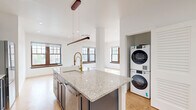 Located on the sixth floor of 250 Harrison Street in downtown Syracuse, NY, this 2-bedroom, 2-bathroom residence offers a well-appointed layout with modern finishes throughout. Cork flooring runs consistently across the living spaces, providing a warm, natural aesthetic underfoot.

The open kitchen is a standout feature, showcasing navy blue shaker cabinetry with gold hardware, granite countertops, a large center island with an undermount sink and a bold brass faucet, stainless steel appliances, and a subway tile backsplash. A sleek linear pendant light anchors the space. The kitchen flows directly into the dining area, which is bathed in natural light from large, wood-framed multi-pane windows overlooking the Syracuse skyline.

Both bedrooms are generously proportioned with clean white walls, cork flooring, and large windows that frame city views. The primary bedroom features multiple windows, filling the room with ample natural light.

The bathrooms are elegantly finished with white subway tile accented by a distinctive green tile border. One bathroom includes a glass-enclosed walk-in shower, while both feature granite-topped vanities with undermount sinks and framed mirrors. A dedicated closet provides practical storage. Recessed lighting throughout the residence adds a polished, contemporary feel.