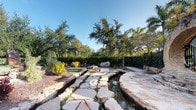 This thoughtfully designed outdoor space showcases a striking natural stone pathway that meanders across a shallow, tranquil water channel, creating a visually engaging and functional landscape feature. The irregularly shaped flagstones are carefully arranged to form a seamless crossing, flanked on both sides by carefully placed boulders and natural stonework that frame the water element with an organic, earthy aesthetic.

The surrounding landscape is lush and well-maintained, featuring a diverse mix of tropical plantings including vibrant bromeliads, ornamental grasses, and mature shade trees that provide a verdant canopy overhead. Stately palm trees punctuate the background, reinforcing the tropical character of the space. Dense, manicured hedgerows line the perimeter, offering privacy and a defined boundary, while a black wrought iron fence provides additional security without compromising the open, airy feel of the environment.

Generous mulched planting beds enrich the overall composition, and the entire space benefits from abundant natural light under clear blue skies. The combination of water, stone, and lush vegetation creates a serene, resort-like ambiance that is both visually captivating and inviting.