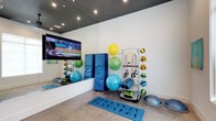 This single-floor fitness studio is thoughtfully designed to support a wide range of exercise routines. A full-length wall mirror spans one side of the room, creating an expansive feel while allowing users to monitor their form during workouts. Recessed lighting set against a dark ceiling provides focused, even illumination throughout the space.

The studio is stocked with an impressive array of functional fitness equipment, including stability balls in multiple sizes, medicine balls, resistance bands, foam rollers, and BOSU balance trainers. A freestanding multi-tier equipment rack keeps accessories neatly organized and easily accessible. Blue folding gymnastics mats offer cushioned support for floor-based exercises and stretching routines. Exercise mats are also available for individual use on the polished concrete-style flooring.

A large wall-mounted flat-screen television is positioned prominently for entertainment or instructional use during sessions. The clean, white walls contribute to a bright and open atmosphere, while the dark ceiling adds a modern, polished contrast. The overall layout is open and uncluttered, providing ample floor space for movement-based workouts, group classes, or individual training sessions.
