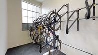 This single-floor bicycle storage room is purpose-built to accommodate cyclists with a practical and well-organized layout. The room features a combination of wall-mounted horizontal rack systems and freestanding vertical bike stands, allowing multiple bicycles to be stored efficiently without occupying unnecessary floor space. The vertical rack configuration enables bikes to be stacked at varying heights, maximizing the room's capacity. A repair stand is also present, providing a convenient station for bicycle maintenance and tune-ups directly within the storage area. The flooring is finished in a dark, durable surface suited for heavy use and easy cleaning. Natural light filters into the room through frosted industrial-style grid windows, lending the space a bright and functional atmosphere. The walls are finished in a clean, neutral tone, contributing to an uncluttered environment. A solid white entry door with a secure lock provides controlled access to the room, ensuring the safety of stored bicycles. Overall, this space is thoughtfully designed to serve cyclists with a secure, accessible, and well-equipped environment for both storing and maintaining their bikes.