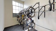 This single-floor bicycle storage room is purpose-built to accommodate cyclists with a practical and well-organized layout. The room features a combination of wall-mounted horizontal rack systems and freestanding vertical bike stands, allowing multiple bicycles to be stored efficiently without occupying unnecessary floor space. The vertical rack configuration enables bikes to be stacked at varying heights, maximizing the room's capacity. A repair stand is also present, providing a convenient station for bicycle maintenance and tune-ups directly within the storage area. The flooring is finished in a dark, durable surface suited for heavy use and easy cleaning. Natural light filters into the room through frosted industrial-style grid windows, lending the space a bright and functional atmosphere. The walls are finished in a clean, neutral tone, contributing to an uncluttered environment. A solid white entry door with a secure lock provides controlled access to the room, ensuring the safety of stored bicycles. Overall, this space is thoughtfully designed to serve cyclists with a secure, accessible, and well-equipped environment for both storing and maintaining their bikes.