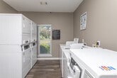 This shared laundry facility is a practical, single-floor amenity space designed to serve a residential community. The room features a clean, neutral color palette with warm gray walls and durable wood-look flooring throughout, creating a tidy and functional environment.

The laundry area is outfitted with a row of commercial-grade top-load and front-load washing machines, complemented by stacked dryer units positioned along the opposite wall. Instructional signage for both washing and drying is prominently displayed, and bilingual CSC service information is posted for resident convenience. A vending machine is also available within the space.

A light wood folding table with seating provides a practical surface for sorting and folding laundry. Natural light filters in through a window with blinds, adding brightness to the room. Overhead flush-mount lighting and a security camera ensure the space remains well-lit and monitored.

A doorway connects the laundry room to an adjacent fitness area, visible through the opening and featuring cardio equipment, further enhancing the utility of this amenity space. The overall layout is efficient and straightforward, offering residents a comfortable and well-maintained space to manage their laundry needs.