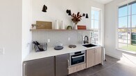 This contemporary single-floor space is thoughtfully designed with clean lines, a neutral white palette, and modern finishes throughout. The interior features large-format gray tile flooring that anchors the open layout, while crisp white walls provide a bright, airy atmosphere. Abundant natural light streams in through grid-pane windows and a glass-paneled door that opens directly onto the outdoor terrace, blurring the boundary between interior and exterior.

The kitchenette is a standout feature, equipped with light wood and stainless cabinetry, a white quartz countertop, a geometric white tile backsplash, a black undermount sink, and a built-in Bosch oven. Open wood shelving above the counter, accented by matte black wall sconces, adds both functional storage and modern style.

The space connects seamlessly to a generous rooftop terrace that offers multiple distinct zones: an artificial turf lawn area, a paved section with café-style seating, a long communal dining table, and a full outdoor grill station. Greenery hedging lines the perimeter for privacy, while overhead string lights create an inviting ambiance. Open sky views and surrounding treetops complete the outdoor experience.