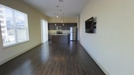 This single-floor space features an open-concept layout that seamlessly connects the living area to the kitchen, creating a spacious and cohesive environment. Dark hardwood floors run throughout, providing a rich, warm contrast to the light-toned walls and adding a sense of depth to the space.

The kitchen is outfitted with dark espresso cabinetry that extends to the upper and lower walls, offering ample storage. Stainless steel appliances, including a refrigerator, microwave, and range, lend a modern, polished look. A central island with a built-in sink serves as a functional focal point, ideal for meal preparation or casual dining. Pendant lighting hangs above the island, providing targeted illumination and a stylish design touch.

The adjacent living area is open and unobstructed, allowing for flexible furniture arrangement. A wall-mounted flat-screen television is positioned on one side, while a large window fitted with vertical blinds allows natural light to filter into the space. The overall ambiance is contemporary and clean, with a neutral color palette and cohesive design elements that create a practical yet inviting atmosphere.