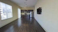 This single-floor space features an open-concept layout that seamlessly connects the living area to the kitchen, creating a spacious and cohesive environment. Dark hardwood floors run throughout, providing a rich, warm contrast to the light-toned walls and adding a sense of depth to the space.

The kitchen is outfitted with dark espresso cabinetry that extends to the upper and lower walls, offering ample storage. Stainless steel appliances, including a refrigerator, microwave, and range, lend a modern, polished look. A central island with a built-in sink serves as a functional focal point, ideal for meal preparation or casual dining. Pendant lighting hangs above the island, providing targeted illumination and a stylish design touch.

The adjacent living area is open and unobstructed, allowing for flexible furniture arrangement. A wall-mounted flat-screen television is positioned on one side, while a large window fitted with vertical blinds allows natural light to filter into the space. The overall ambiance is contemporary and clean, with a neutral color palette and cohesive design elements that create a practical yet inviting atmosphere.