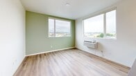 Located on the fifth floor at 222 Capitol Way N in downtown Olympia, Washington, this single-floor residence features a clean, contemporary design throughout. Light wood-tone flooring runs continuously across the space, lending warmth and visual cohesion to each area.

The kitchen is well-equipped with white shaker cabinetry extending to the upper walls, gray countertops, a stainless steel sink, and a full suite of stainless steel appliances including a refrigerator, electric range, and dishwasher. Recessed lighting and ample cabinet storage make the kitchen both functional and visually appealing.

The living area is generously proportioned, with large windows on two walls that frame elevated views of the surrounding Olympia cityscape and tree canopy. A distinctive sage green accent wall adds a pop of color, while a wall-mounted HVAC unit ensures year-round comfort.

Additional rooms include a bedroom with a sliding closet door and a window offering city views, as well as a second versatile room with its own window. A hallway connects the spaces efficiently.

The bathroom features white cabinetry, a gray-topped vanity, and a well-lit mirror. A stacked in-unit washer and dryer is conveniently located nearby, adding practical functionality to this thoughtfully designed residence.