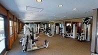This fitness center offers a thoroughly equipped workout environment designed to support a wide range of exercise routines. The space is anchored by a generous floor plan that accommodates an extensive array of StairMaster strength training machines and selectorized equipment, arranged in an open layout that allows for comfortable movement between stations. Recessed lighting throughout the ceiling provides consistent, even illumination across the entire floor, enhancing visibility and creating a clean, professional atmosphere. Large mirrored wall panels line the perimeter, visually expanding the space while allowing users to monitor their form during workouts. The neutral-toned carpeted flooring adds a layer of comfort underfoot and contributes to the room's polished, well-maintained appearance. Cardio equipment, including stair climbers, is positioned toward the back of the space, rounding out the full-body fitness offering. The overall ambiance is functional yet refined, reflecting a facility built to meet the needs of dedicated fitness enthusiasts in a comfortable, organized setting.