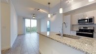This single-floor residence offers a seamlessly connected open-plan layout that integrates the kitchen and living areas into one cohesive, light-filled space. The kitchen is anchored by a generously sized island topped with speckled granite countertops, complemented by a polished chrome faucet and an undermount sink. White cabinetry lines the kitchen perimeter, contributing to the clean, contemporary aesthetic throughout. Pendant lighting fixtures hang above the island, while a track lighting system illuminates the broader living area, together creating a layered and functional lighting scheme. The living space features light-toned large-format tile flooring, adding to the airy, modern feel. A ceiling fan is centrally positioned, enhancing comfort and airflow. Floor-to-ceiling sliding glass doors flood the interior with natural light and provide direct access to a private balcony enclosed by a metal railing, offering pleasant views of surrounding greenery. The overall ambiance is bright, clean, and contemporary, with an emphasis on open space and natural light throughout.
