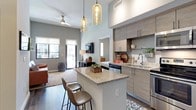 This two-bedroom, two-bathroom residence offers a thoughtfully designed open-concept layout that seamlessly connects the kitchen, dining, and living areas. Soaring ceilings amplify the sense of space throughout, while dark hardwood-style flooring grounds the contemporary aesthetic.

The kitchen is a standout feature, equipped with stainless steel appliances including a range, microwave, dishwasher, and refrigerator. Granite countertops extend across a central island with bar seating for two, illuminated by a pair of pendant globe lights overhead. Taupe wood-grain cabinetry provides ample storage, complemented by open shelving for display.

The living area benefits from large windows and direct access to a private outdoor space, inviting natural light throughout the day. A ceiling fan enhances comfort, and wall-mounted planters add a natural, organic touch to the neutral gray walls.

Both bathrooms are finished with granite vanity tops, wood-toned cabinetry, framed mirrors, and tiled tub-shower combinations. The bedrooms, visible from the main living space, offer a quiet retreat with direct sightlines kept minimal for privacy. The overall ambiance is modern, clean, and well-appointed.