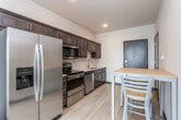 This single-floor unit offers a well-organized, functional layout designed to maximize comfort and convenience. The kitchen is a standout feature, outfitted with a full suite of stainless steel appliances including a refrigerator, electric range, built-in microwave, and dishwasher. Rich dark-brown shaker-style cabinetry lines the walls, complemented by light-toned countertops and a stainless steel sink. Light wood-style flooring runs throughout, lending a clean and cohesive aesthetic to the space.

Adjacent to the kitchen, the living area provides a welcoming atmosphere, benefiting from natural light through a large window fitted with vertical blinds. The bedroom is compact yet practical, featuring a window with an exterior view and a bed frame with integrated drawer storage underneath.

A walk-in closet with wire shelving provides ample storage, positioned conveniently near the bathroom, which includes a dark-cabinetry vanity with a large mirror and a full soaking tub with shower. A dedicated in-unit laundry room houses a full-sized washer and dryer beneath additional wire shelving, completing this efficiently designed and move-in-ready living space.