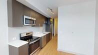 This single-floor space showcases a clean, modern design with thoughtful finishes throughout. The kitchen is a standout feature, boasting a galley-style layout with rich dark wood cabinetry that extends to the ceiling, providing generous storage space. Contrasting white countertops create a striking visual balance, while a subway tile backsplash adds a refined, contemporary touch. The kitchen is fully equipped with a complete suite of stainless steel appliances, including an electric range, built-in microwave, dishwasher, and refrigerator. A sleek undermount sink with a modern arc faucet anchors the countertop workspace. Track lighting mounted on the ceiling ensures excellent task illumination throughout the kitchen. Light-toned hardwood flooring flows seamlessly from the kitchen into the adjacent hallway and living areas, adding warmth and continuity to the overall aesthetic. The walls are finished in crisp white, enhancing the sense of brightness and openness. The layout is efficient and well-organized, with a corridor leading further into the unit. The overall ambiance is contemporary and polished, offering a clean and functional living environment with a cohesive design palette of dark wood, white surfaces, and stainless steel accents.