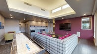 This dynamic single-floor amenity space is thoughtfully designed to accommodate both socializing and productive gatherings. The central lounge area is anchored by a generously sized plaid sectional sofa accented with bold magenta throw pillows, complemented by a shuffleboard table and a wall-mounted flat-screen television set against a striking deep-magenta accent wall. Soaring clerestory windows and a raised ceiling coffer flood the lounge with abundant natural light, creating an open and inviting atmosphere.

Adjacent to the lounge, a casual seating nook features bright yellow armchairs alongside a classic Pac-Man arcade cabinet and colorful wall art, adding a playful, retro touch. A high-top bar counter with pendant lighting and city views provides an ideal spot for casual dining or socializing.

The fully equipped kitchen is a standout feature, boasting stone countertops, light wood and gray cabinetry, subway tile backsplash, stainless steel appliances including a gas range and built-in microwave, and a large island with seating for multiple guests — all illuminated by industrial-style pendant lights.

A dedicated conference and dining area offers a long communal table surrounded by modern white chairs, bathed in natural light from wraparound clerestory windows and direct access to an outdoor terrace. The terrace extends the entertaining space with urban skyline views, completing this well-appointed community gathering destination.