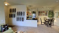 This single-floor residence offers a thoughtfully designed open-concept layout that connects the living, dining, and kitchen areas into one cohesive and functional space. The living area features plush wall-to-wall carpeting and is accented by decorative wall art, adding a touch of personality and warmth to the space. A half-wall breakfast bar elegantly divides the kitchen from the main living area, providing both a visual boundary and a casual dining surface. The kitchen is equipped with modern stainless steel appliances, including a refrigerator and range, complemented by warm wood-toned cabinetry and dark countertops. Track lighting runs along the ceiling in both the kitchen and adjoining areas, ensuring bright, well-distributed illumination throughout. The dining area sits adjacent to the kitchen and living space, featuring a glass-top dining table with stylish chairs, making it ideal for everyday meals or entertaining. The neutral color palette of warm beige walls and light carpeting creates a clean, inviting ambiance that flows naturally from room to room, making the overall space feel open, airy, and easy to personalize.