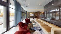 This elegantly designed single-floor amenity space combines modern luxury with functional versatility. The main lounge area is anchored by a dramatic dark marble accent wall mounted with a large flat-screen television, flanked by generous caramel-toned sectional sofas and bold crimson velvet armchairs arranged atop artfully patterned area rugs. An ornate metallic room divider adds a sculptural element, separating the lounge from a billiards area beyond.

Floor-to-ceiling black-framed glass walls and windows line the perimeter, bathing the interior in abundant natural light while offering open views to the outdoors. The expansive layout flows seamlessly from the social seating areas into a well-appointed kitchenette, which features rich walnut cabinetry, stainless steel appliances including a French door refrigerator, a striking stone-topped island, open display shelving, and distinctive pendant lighting. A large abstract artwork adds a vibrant artistic focal point near the kitchen.

Recessed lighting throughout the space complements the natural light, while light-toned large-format tile flooring grounds the design with a clean, polished aesthetic. The overall ambiance is one of refined, contemporary sophistication, making the space well-suited for both relaxed gatherings and elevated social events.