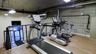 This two-floor fitness facility offers a well-equipped environment designed for effective workouts. The space features a selection of professional-grade True Fitness cardio machines, including treadmills, ellipticals, and a stationary bike, all positioned on clean hardwood-style flooring that adds warmth to the utilitarian setting. The interior showcases an industrial aesthetic, with exposed concrete block walls, visible ductwork, and overhead piping that contribute to a raw, functional atmosphere. Overhead lighting illuminates the workout area efficiently, ensuring visibility across the floor. A railing system separates sections of the space, suggesting a mezzanine or multi-level layout that maximizes the available square footage across both floors. The open layout allows for easy movement between machines and provides a practical, no-frills environment focused on fitness. The combination of durable surfaces and sturdy equipment reflects a space built for consistent, heavy use — making it well-suited for a commercial, residential amenity, or institutional fitness setting.