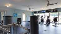 This fitness center is a single-floor facility designed to support a well-rounded workout experience. The space features soft blue walls, a dark rubberized flooring surface, and ample natural light streaming in through large windows along one wall, creating an open and energizing atmosphere. Ceiling fans are positioned throughout the room to ensure comfortable airflow during workouts.

The cardio section is lined with elliptical trainers and treadmills positioned directly in front of the windows, allowing users to enjoy outdoor views while exercising. A row of flat-screen televisions is mounted along the upper portion of the wall above the cardio equipment, providing entertainment options during sessions. Track lighting supplements the natural light in this area.

On the opposite side of the room, a dedicated free weights area includes a multi-tier dumbbell rack with a full range of weights, accompanied by a weight bench for strength training exercises. Informational boards are posted on the walls, offering guidance and resources for facility users.

The space is accessible via a door at the rear, and its efficient layout ensures that both cardio and strength training zones are clearly defined without feeling cramped. Overall, the fitness center presents a clean, functional, and well-maintained environment suited for a variety of exercise routines.