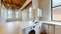 Located at 425 W Lakeside Ave in Cleveland, OH, this one-bedroom, one-bathroom loft showcases a compelling blend of historic industrial character and modern finishes. The open-concept kitchen and living area serves as the heart of the unit, where whitewashed exposed brick walls meet striking wood beam ceilings—a hallmark of the building's original construction. Light hardwood flooring runs throughout, enhancing the bright, airy atmosphere created by multiple black-framed windows that frame urban city views.

The kitchen is thoughtfully designed with white shaker-style cabinetry, white quartz countertops, a subway tile backsplash, and stainless steel appliances including a dishwasher and range. An in-unit LG washer is seamlessly integrated beneath the countertop for added convenience.

The bedroom offers a comfortable, well-proportioned layout with dual windows that allow natural light and city views to fill the space. Two closets provide ample storage throughout the unit.

The bathroom features clean, modern finishes including a white vanity with black hardware accents, a framed mirror, and hardwood-style flooring that ties the space together cohesively. A hallway connects the main living areas, completing this well-appointed urban loft.