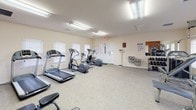 This fitness center is a thoughtfully designed, single-floor facility built to support a comprehensive workout experience. The space is divided into distinct functional zones, seamlessly flowing from one to the next across carpeted flooring that adds comfort underfoot.

The cardio area is lined with multiple treadmills, elliptical trainers, and a stationary recumbent bike, all positioned alongside a row of windows fitted with blinds that allow natural light to fill the room while maintaining privacy. Overhead fluorescent lighting ensures consistent, bright illumination throughout.

The strength training zone features a multi-station cable machine, a functional trainer, and various benches for free weight exercises. A dumbbell rack and instructional fitness charts posted on the walls make the space practical and user-friendly. An accent wall painted in a bold burnt orange provides a striking visual focal point, adorned with motivational words including "Motivation," "Strength," and "Imagine," reinforcing an energizing atmosphere.

The overall ambiance is clean and purposeful, with light-colored walls complementing the vibrant accent wall to create a balanced, welcoming environment. An emergency exit and a secondary door offer convenient access throughout the space. This fitness center is well-suited for a broad range of workout routines, from cardio sessions to full-body strength training.