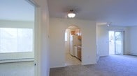 This single-floor residential space offers a straightforward and functional layout designed for comfortable everyday living. The living area is finished with wall-to-wall carpeting that adds warmth underfoot, while smooth white walls and a ceiling fan with a light fixture provide a clean, bright atmosphere. A distinctive arched entryway serves as the defining architectural detail, elegantly connecting the living area to the kitchen and adding a touch of classic charm to the otherwise simple design. The kitchen is outfitted with warm wood-toned cabinetry, white appliances including a refrigerator and dishwasher, and light-toned countertops. A window in the kitchen allows natural light to filter into the space, keeping the area feeling open and airy. The transition between the carpeted living zone and the hard-surface kitchen flooring is seamless through the arched passage. Additional windows visible from the living area further enhance the sense of light and openness throughout. The overall ambiance of the space is practical and welcoming, with a neutral color palette that provides a versatile backdrop suitable for a variety of furnishing styles.