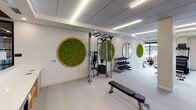 This thoughtfully designed fitness center offers a comprehensive workout environment spread across a single level. The space is divided into distinct functional zones, each tailored to a specific type of training. The cardio area is lined with floor-to-ceiling glass windows that flood the room with natural light and provide open outdoor views, housing a full complement of treadmills, stationary bikes, ellipticals, and stair climbers. A dedicated strength training zone features a multi-station cable machine, a tiered dumbbell rack, and adjustable benches, providing ample options for resistance workouts. A standout feature is the glass-enclosed studio, which contains a cycling bike, wall-mounted display, and a ballet barre, offering a private setting for focused or instructor-led sessions. Throughout the main area, circular framed moss wall installations bring a biophilic design element that adds warmth and visual interest to the otherwise clean, neutral palette. Light wood-toned flooring, a drop ceiling with recessed and linear LED lighting, and crisp white walls contribute to a polished, contemporary ambiance. A vertical slatted wood accent wall near the studio entrance adds an additional layer of refined detailing.