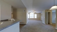 This single-story space offers an open and airy layout highlighted by vaulted ceilings that rise dramatically in the main living area, lending a sense of generous volume and grandeur. A ceiling fan is centrally positioned beneath the peak of the vault, providing both function and a subtle architectural focal point. The living area is carpeted in a neutral tone, complementing the warm beige walls throughout and contributing to a cohesive, understated palette.

Natural light flows generously into the space through a prominent arched window flanked by additional rectangular windows at the far end of the living area. This window arrangement not only brightens the interior but also serves as a visually striking architectural feature. White baseboards frame the perimeter of the room, adding a clean, finished look.

Adjacent to the living area, the kitchen is partially visible and features granite-style countertops with a built-in sink, offering a functional and attractive workspace. The open sightlines between the kitchen and living space encourage a connected, social atmosphere. Overall, the space presents a bright, comfortable, and well-designed environment suitable for everyday living.