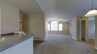 This single-story space offers an open and airy layout highlighted by vaulted ceilings that rise dramatically in the main living area, lending a sense of generous volume and grandeur. A ceiling fan is centrally positioned beneath the peak of the vault, providing both function and a subtle architectural focal point. The living area is carpeted in a neutral tone, complementing the warm beige walls throughout and contributing to a cohesive, understated palette.

Natural light flows generously into the space through a prominent arched window flanked by additional rectangular windows at the far end of the living area. This window arrangement not only brightens the interior but also serves as a visually striking architectural feature. White baseboards frame the perimeter of the room, adding a clean, finished look.

Adjacent to the living area, the kitchen is partially visible and features granite-style countertops with a built-in sink, offering a functional and attractive workspace. The open sightlines between the kitchen and living space encourage a connected, social atmosphere. Overall, the space presents a bright, comfortable, and well-designed environment suitable for everyday living.