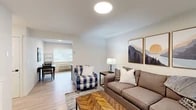 Located at 1000 W Ironwood Dr in Coeur d'Alene, Idaho, this well-appointed single-floor unit offers a clean, contemporary design with warm, nature-inspired accents throughout. The open living area serves as the heart of the space, featuring light wood-look flooring, a wall-mounted electric fireplace, and a flat-screen TV. A herringbone-patterned wood coffee table and a mix of plaid and neutral upholstered seating add texture and character, while mountain-themed artwork echoes the natural beauty of the Pacific Northwest. A teal front door and bright windows with slatted blinds allow natural light to fill the room. The dining area flows seamlessly from the living space, providing a functional area for meals and gatherings. The kitchen is equipped with stainless steel appliances including a range, microwave, and dishwasher, complemented by two-tone cabinetry — warm wood upper cabinets with open shelving and bold blue lower cabinets — set against crisp white countertops. The primary bedroom offers a comfortable retreat with a large window, light flooring, and ample closet space with sliding doors. A second bedroom functions well as a guest room or home office, featuring a desk, chair, and natural light. The bathroom presents a clean, neutral palette with a full tub-shower combination, gray vanity, and white countertop.