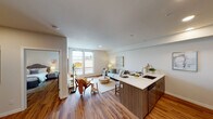 This thoughtfully designed single-floor residence offers a seamless blend of modern aesthetics and functional living. The open-concept layout connects the kitchen and living area, creating a bright and welcoming communal space. Warm hardwood-style flooring flows throughout, anchoring the neutral color palette of soft whites and greiges that lend the unit a clean, contemporary feel.

The kitchen serves as a central hub, featuring a large peninsula with a white quartz countertop that provides ample workspace and doubles as a casual dining area. Walnut-toned cabinetry pairs with stainless steel appliances — including a refrigerator, electric range, and built-in microwave — while recessed lighting overhead ensures the space is well-lit. A bold blue entry door adds a distinctive design accent.

The living area is framed by large windows that draw in generous natural light and offer exterior views, giving the space an open, airy atmosphere. The bedroom continues the warm, neutral tone with hardwood flooring, a bright window overlooking the surrounding neighborhood, and tasteful wall art that contributes to the room's calm ambiance.

The bathroom is finished with a wood-grain vanity, a white quartz countertop, a large mirror with overhead lighting, and a full soaking tub with a shower — all finished in a clean, understated style consistent with the rest of the unit.