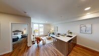 This thoughtfully designed single-floor residence offers a seamless blend of modern aesthetics and functional living. The open-concept layout connects the kitchen and living area, creating a bright and welcoming communal space. Warm hardwood-style flooring flows throughout, anchoring the neutral color palette of soft whites and greiges that lend the unit a clean, contemporary feel.

The kitchen serves as a central hub, featuring a large peninsula with a white quartz countertop that provides ample workspace and doubles as a casual dining area. Walnut-toned cabinetry pairs with stainless steel appliances — including a refrigerator, electric range, and built-in microwave — while recessed lighting overhead ensures the space is well-lit. A bold blue entry door adds a distinctive design accent.

The living area is framed by large windows that draw in generous natural light and offer exterior views, giving the space an open, airy atmosphere. The bedroom continues the warm, neutral tone with hardwood flooring, a bright window overlooking the surrounding neighborhood, and tasteful wall art that contributes to the room's calm ambiance.

The bathroom is finished with a wood-grain vanity, a white quartz countertop, a large mirror with overhead lighting, and a full soaking tub with a shower — all finished in a clean, understated style consistent with the rest of the unit.