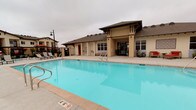 This outdoor amenity space serves as the communal hub of a multi-building residential complex, centered around a generously sized swimming pool with clear, blue water. The pool deck is finished in light-toned, large-format concrete pavers that provide a clean and modern aesthetic while offering ample space for lounging and recreation. Surrounding the pool, a collection of chaise lounge chairs and red market umbrellas are thoughtfully arranged, creating comfortable spots for relaxation and shade.

A decorative focal wall featuring a sunburst medallion design anchors one end of the pool area, adding a distinctive architectural accent to the outdoor space. Adjacent to the pool, a well-designed clubhouse structure with a covered entryway, exposed timber detailing, and a hip roof serves as a community gathering and amenity facility. Its warm, neutral stucco exterior complements the overall Mediterranean-inspired architectural style of the surrounding residential buildings.

The complex buildings, visible in the background, rise two to three stories and feature balconies, warm beige stucco exteriors, and pitched roofs that maintain a cohesive design language throughout. A secured wrought-iron perimeter fence encloses the pool area, ensuring privacy and safety. The space is well-suited for both leisurely relaxation and active outdoor enjoyment.