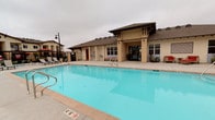 This outdoor amenity space serves as the communal hub of a multi-building residential complex, centered around a generously sized swimming pool with clear, blue water. The pool deck is finished in light-toned, large-format concrete pavers that provide a clean and modern aesthetic while offering ample space for lounging and recreation. Surrounding the pool, a collection of chaise lounge chairs and red market umbrellas are thoughtfully arranged, creating comfortable spots for relaxation and shade.

A decorative focal wall featuring a sunburst medallion design anchors one end of the pool area, adding a distinctive architectural accent to the outdoor space. Adjacent to the pool, a well-designed clubhouse structure with a covered entryway, exposed timber detailing, and a hip roof serves as a community gathering and amenity facility. Its warm, neutral stucco exterior complements the overall Mediterranean-inspired architectural style of the surrounding residential buildings.

The complex buildings, visible in the background, rise two to three stories and feature balconies, warm beige stucco exteriors, and pitched roofs that maintain a cohesive design language throughout. A secured wrought-iron perimeter fence encloses the pool area, ensuring privacy and safety. The space is well-suited for both leisurely relaxation and active outdoor enjoyment.