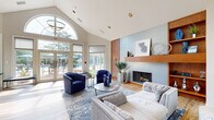 Located at 6959 Castle Ct in Frederick, Maryland, this single-floor community clubhouse is designed to serve as a welcoming and functional gathering space. The main lounge is the architectural centerpiece, boasting soaring vaulted ceilings with a dramatic arched window that floods the room with natural light. A wood-mantled fireplace set against a soft blue accent wall anchors the seating area, which features contemporary gray sofas and bold cobalt blue accent chairs arranged over a patterned area rug atop light hardwood flooring. French doors along the rear wall open directly to an outdoor pool and patio area, seamlessly connecting indoor and outdoor living. A built-in wood bookshelf near the entry adds warmth and character to the space. Adjacent to the lounge, a versatile dining and workspace area includes a large conference-style table, wall-mounted television, and a dedicated desk station with garden views through a glass door. The kitchen is well-appointed with slate-blue cabinetry, a textured brick-style tile backsplash, stainless steel appliances including a full-size refrigerator and microwave, and an expansive light-colored countertop peninsula. The entry foyer features large windows overlooking the exterior walkway, creating a bright and inviting welcome. Throughout, the space maintains a cohesive coastal-contemporary aesthetic with a calming palette of blues, whites, and warm wood tones.