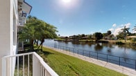 This single-story waterfront property offers a compelling blend of natural beauty and relaxed Florida living. The standout feature of the space is its direct access to a calm, expansive canal, visible from what appears to be a covered porch or deck framed by a classic white railing. The outdoor area extends to a well-maintained lawn that leads to a paved waterside walkway lined with a dark metal fence, creating a clear boundary between the lush green grounds and the shimmering water beyond. Mature tropical trees, including palms and broad-canopied varieties, provide natural shade and enhance the lush, verdant setting. The canal view stretches across to neighboring waterfront homes on the opposite bank, complete with private docks, offering a picturesque and tranquil panorama. Under bright Florida sunshine, the water reflects a vivid blue sky, contributing to the overall sense of openness and natural light that defines the outdoor living experience here. The property's single-floor layout and seamless connection between interior and exterior spaces make it well-suited for comfortable, accessible living. While interior details are limited from the available imagery, the outdoor environment alone speaks to the property's exceptional setting and waterfront character.