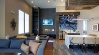 This stylish single-floor space showcases a thoughtfully designed open-concept layout that blends comfort with contemporary aesthetics. The living area is anchored by a striking floor-to-ceiling dark feature wall, housing a linear gas fireplace and a wall-mounted television, creating a dramatic focal point that commands attention. Built-in shelving flanks the feature wall, offering both functional storage and display opportunities.

The lounge area is furnished with plush, sectional seating in rich charcoal tones, accented by warm throw pillows and centered around a solid wood coffee table. Layered area rugs add texture and define the seating space over the hardwood flooring below.

Flowing seamlessly from the living area, the kitchen and dining zone features a clean white countertop island with bar seating, stainless steel appliances, and a textured stone accent wall that adds warmth and visual depth. A large-scale nature-inspired artwork serves as a vibrant backdrop, drawing the eye and infusing the space with an organic element.

Recessed lighting throughout ensures even illumination, while large windows invite natural light. The overall ambiance is polished and modern, blending dark, moody tones with natural textures for a refined and inviting atmosphere.